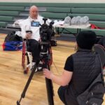Bald man in a wheelchair with fencing gear being interviewed