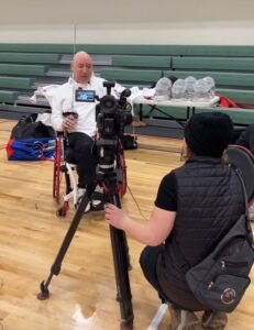 Bald man in a wheelchair with fencing gear being interviewed