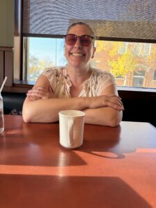 Jenel has dark hair, glasses and a white blouse.She is smiling.